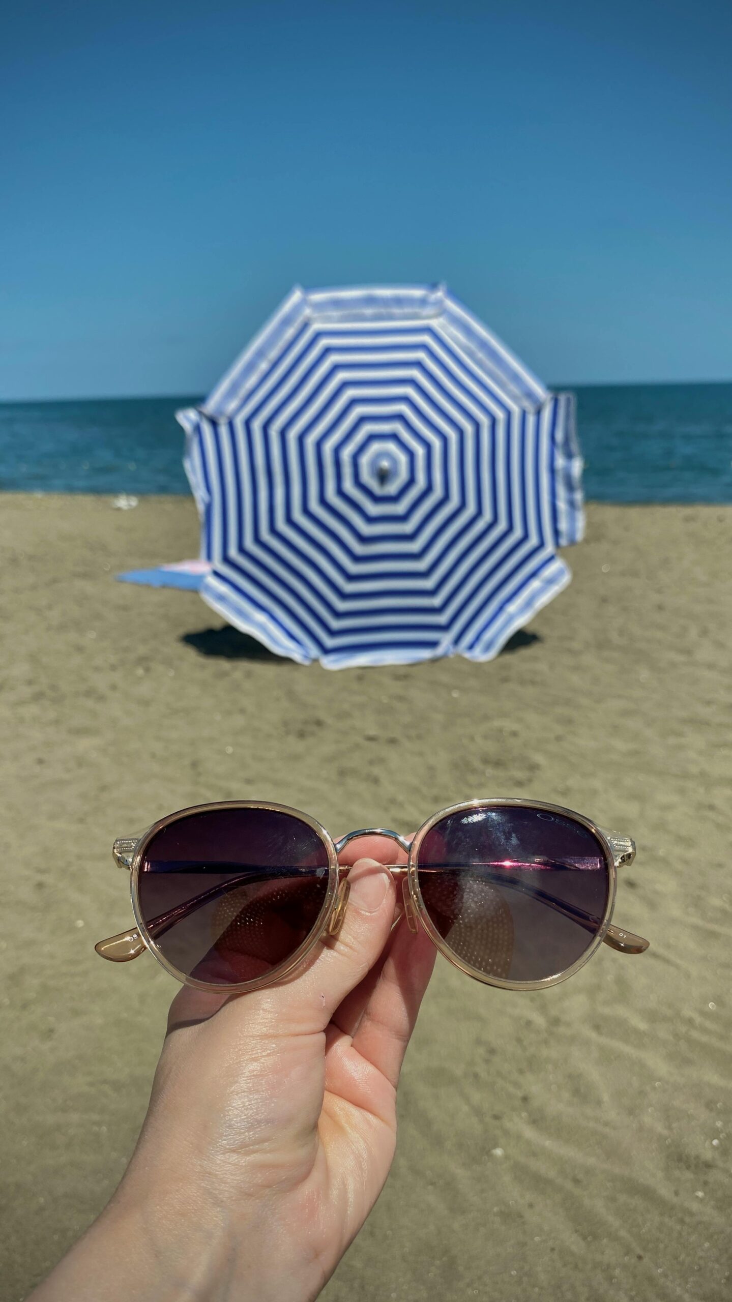 A sunny beach with sunglasses in focus and a blue striped parasol.