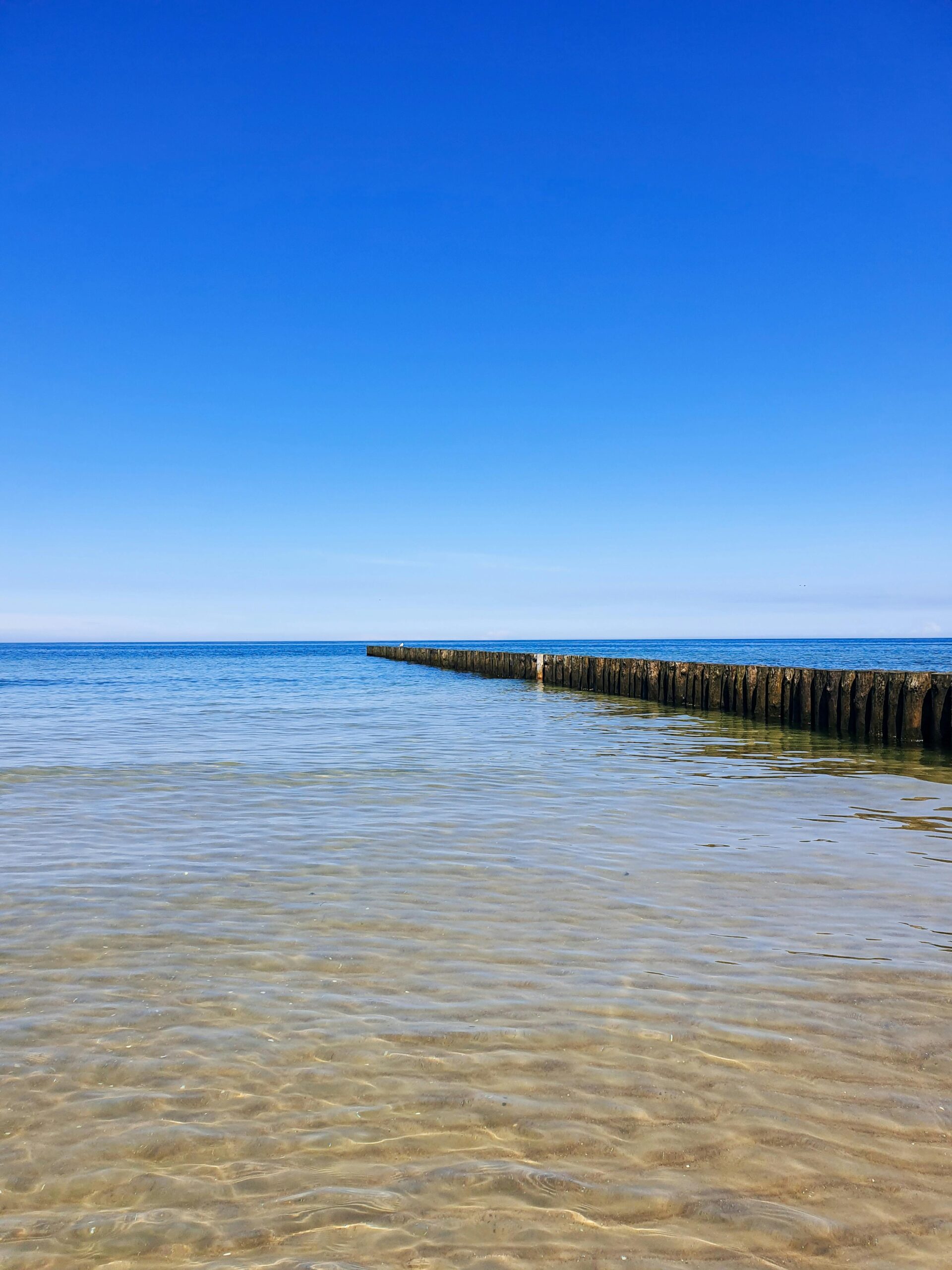 Serene beach scene with a clear blue sky and a wooden pier extending into the calm sea.