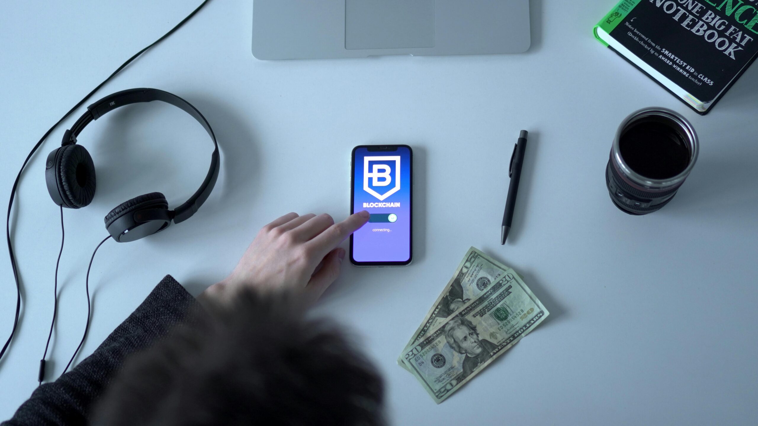 Overhead view of blockchain app on smartphone with cash, headphones, and notebook.